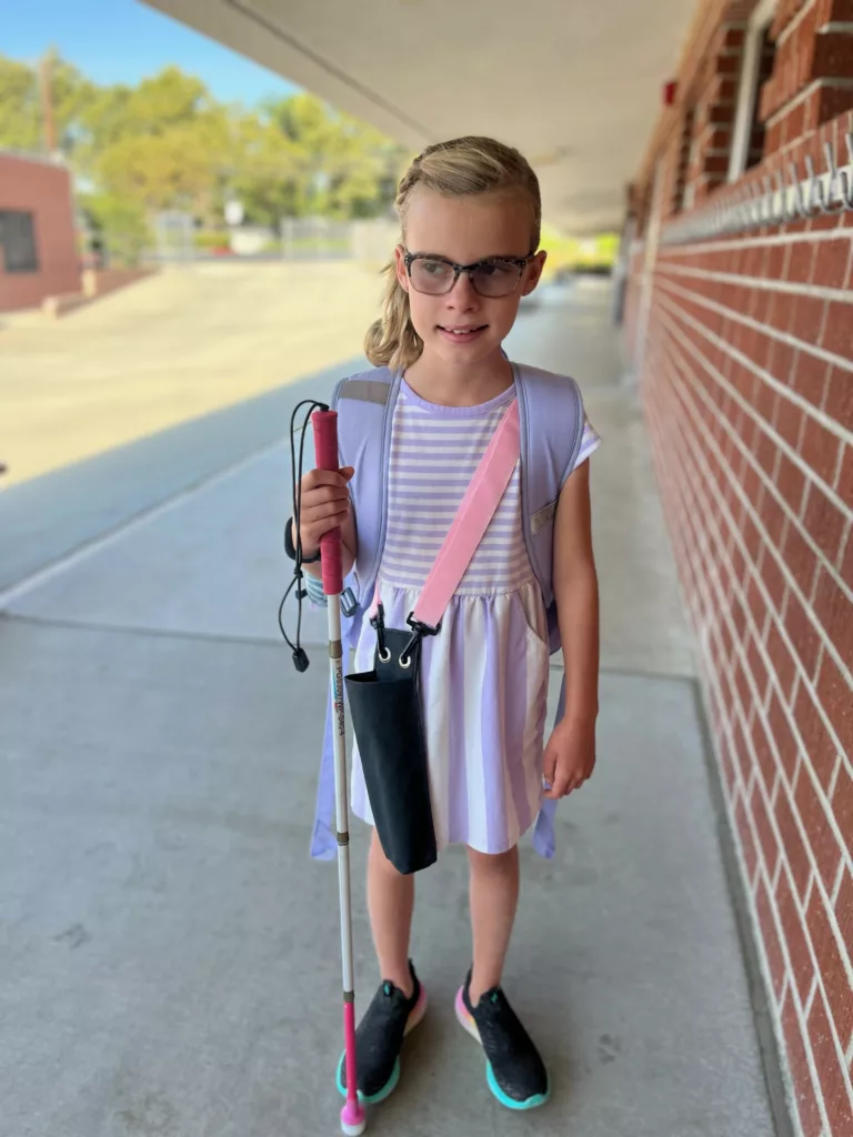 A young girls stands outside her school holding a white cane. 