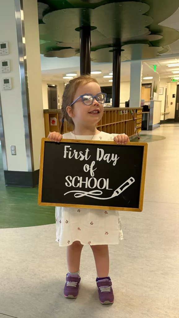 A young girl with glasses standing in school hallway with sign that says "First Day of School"