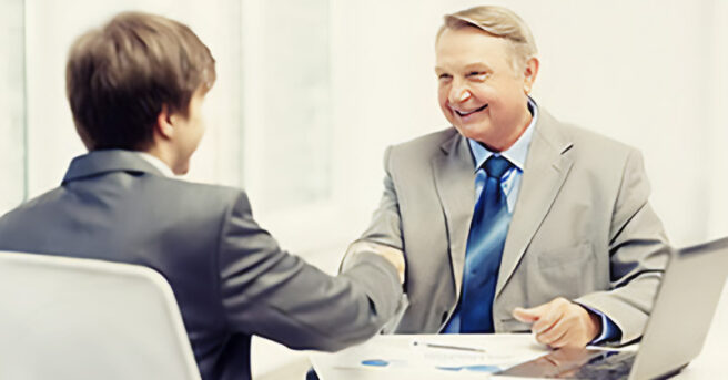 Two people in suits shake hands across a desk