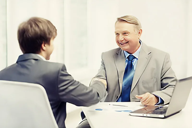 Two people in suits shake hands across a desk