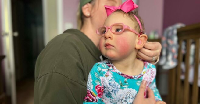 A toddler wearing pink glasses with Mom helping adjust her hearing aid.