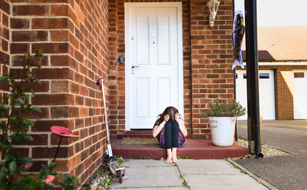 An upset child is sitting on the step in front of their home. The child is holding their head in their hands and resting their forehead on their knees.