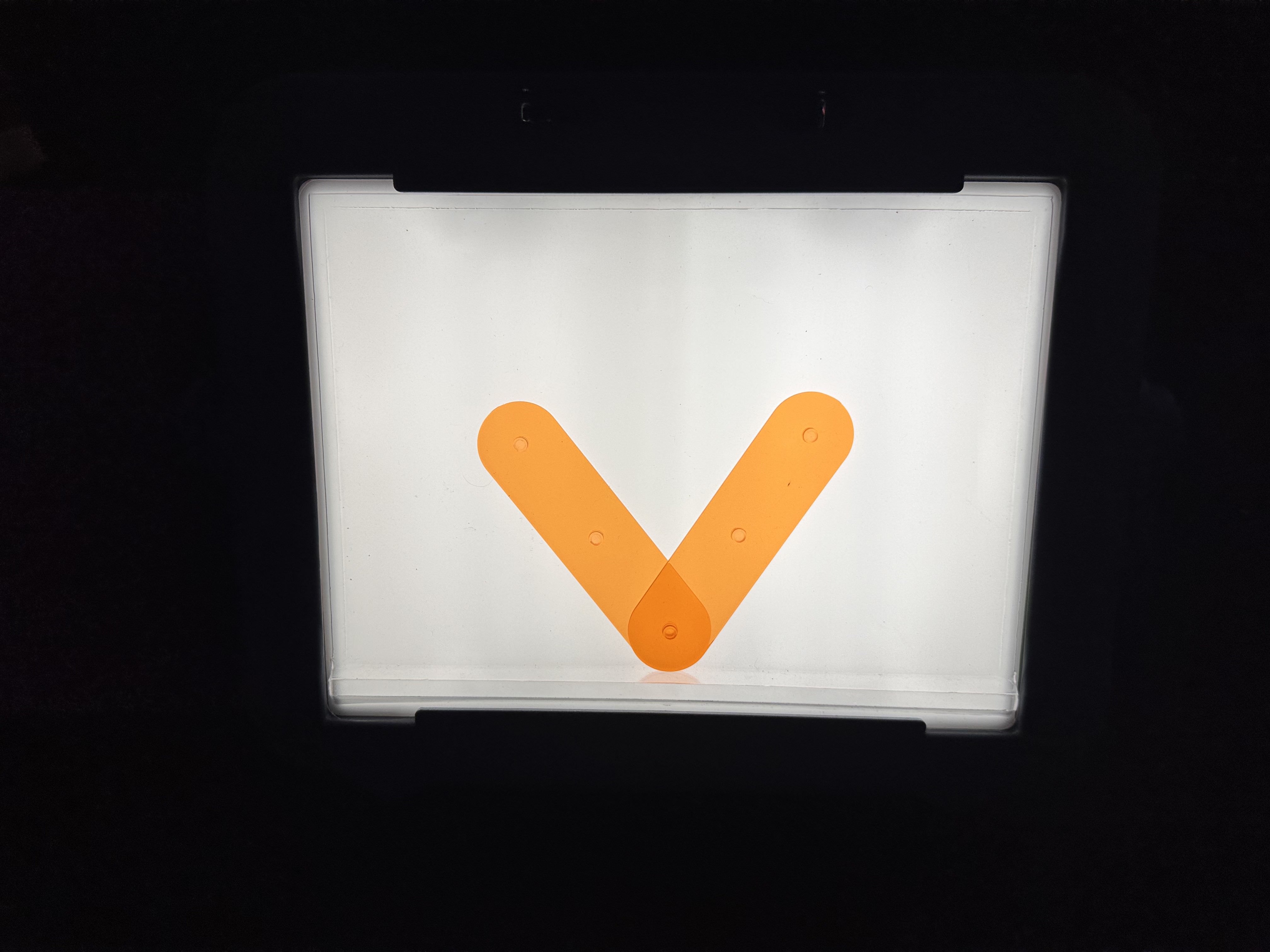 Two transparent, orange lines interlocking to create a lowercase letter “v” on a lightbox in a dark room. 