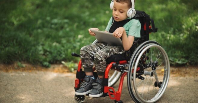 Young child in a wheelchair holding a tablet and wearing headphones.
