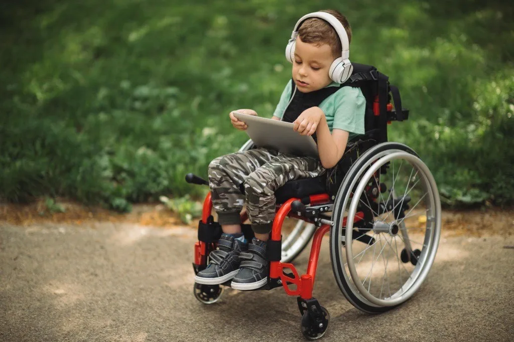 Young child in a wheelchair holding a tablet and wearing headphones.